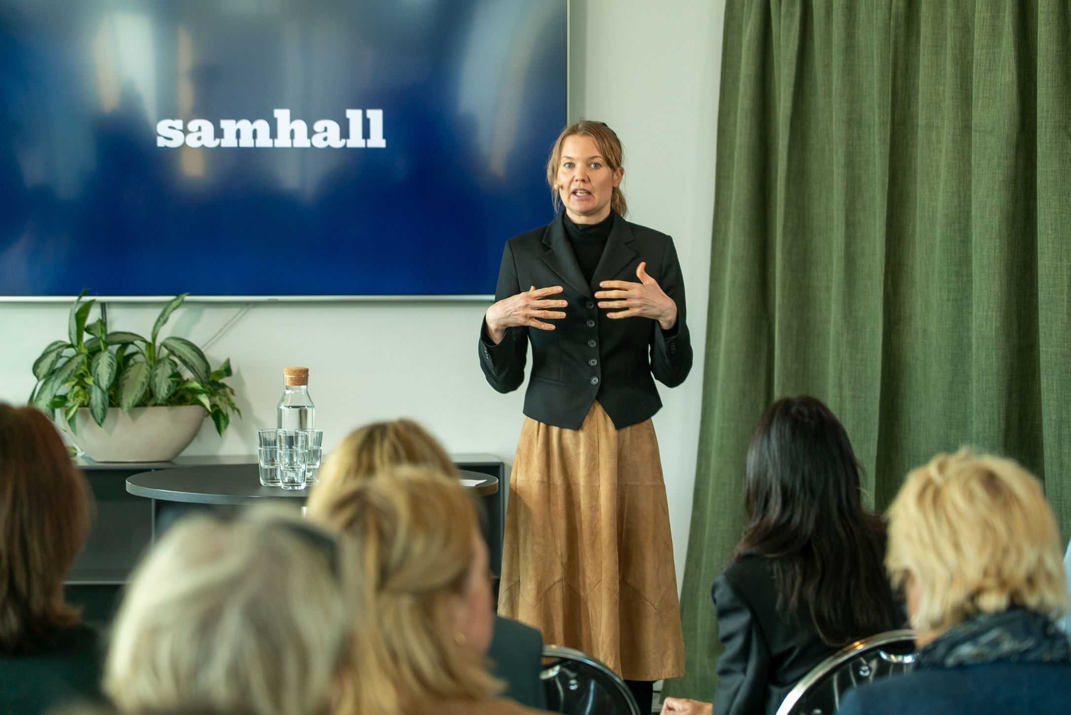 En person står och presenterar inför en sittande publik i ett mötesrum. På en stor skärm bakom personen syns Samhalls logotyp.