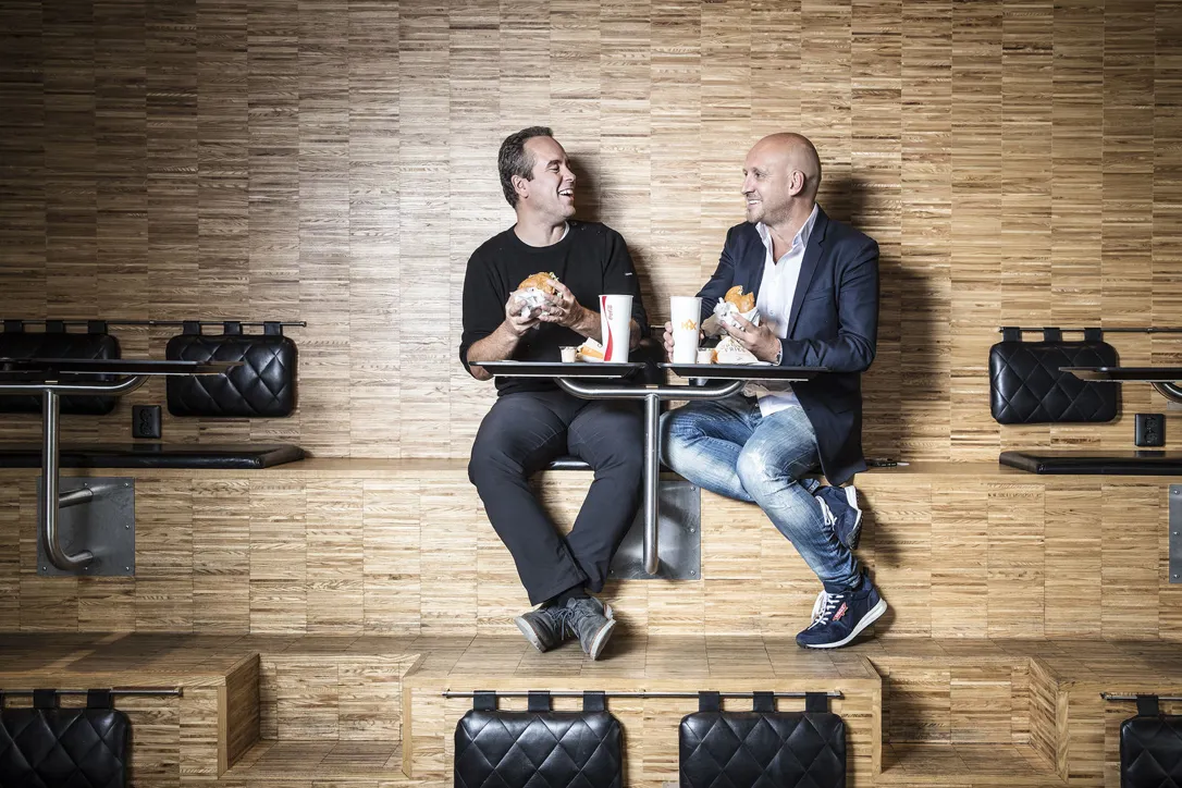 Två män sitter vid ett litet bord mot en träpanelvägg och äter hamburgare medan de skrattar och samtalar. På bordet står pappersmuggar och brickor.